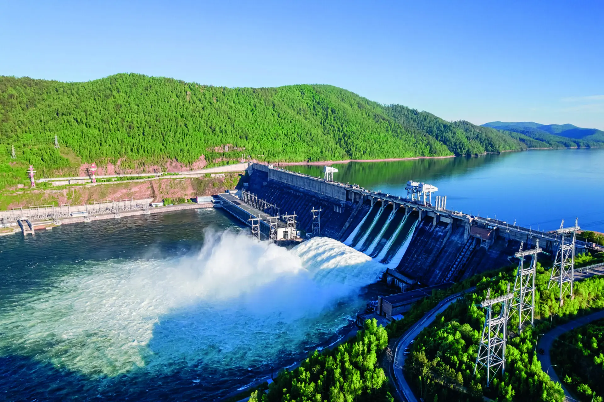 Großes Wasserkraftwerk mit Staudamm, ablaufendem Wasser und umgebenden Wäldern bei klarem Himmel, Luftaufnahme