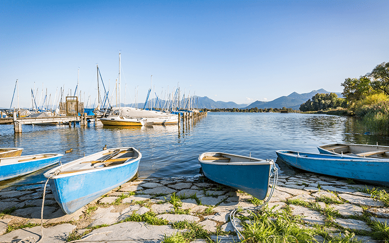 Kleine blaue Ruderboote am Ufer des Chiemsees vor einem Segelhafen mit zahlreichen Segelbooten und Bergpanorama unter klarem Himmel.
