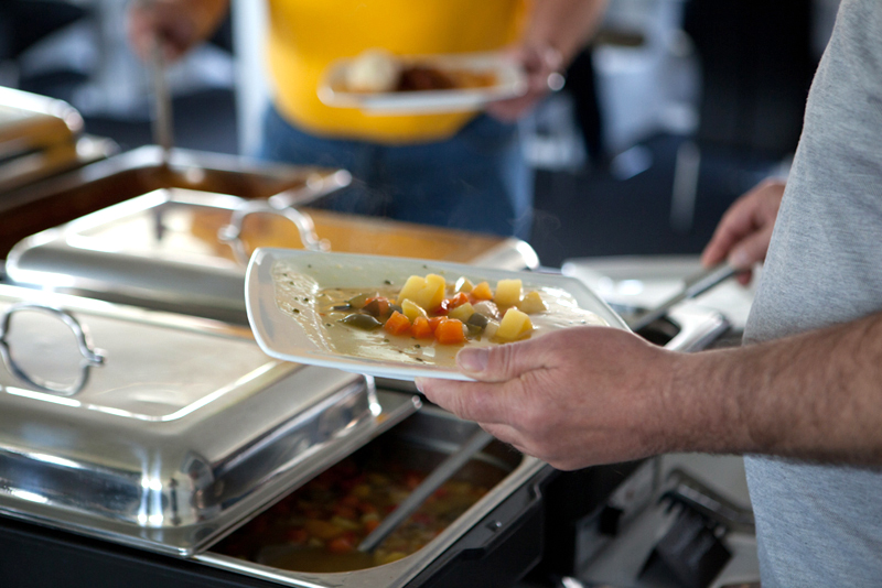 Person schöpft sich an einem warmen Buffet eine Gemüsesuppe auf den Teller, im Vordergrund stehen mehrere Edelstahl-Chafing-Dishes.