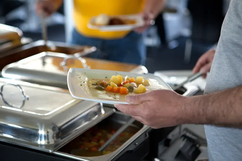 Person schöpft sich an einem warmen Buffet eine Gemüsesuppe auf den Teller, im Vordergrund stehen mehrere Edelstahl-Chafing-Dishes.
