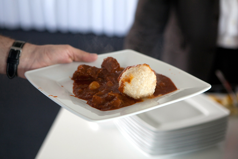 Hand hält einen rechteckigen Teller mit dampfendem Gulasch und einem Kloß, im Hintergrund stehen gestapelte weiße Teller.
