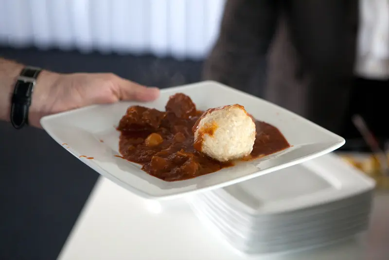 Hand hält einen rechteckigen Teller mit dampfendem Gulasch und einem Kloß, im Hintergrund stehen gestapelte weiße Teller.