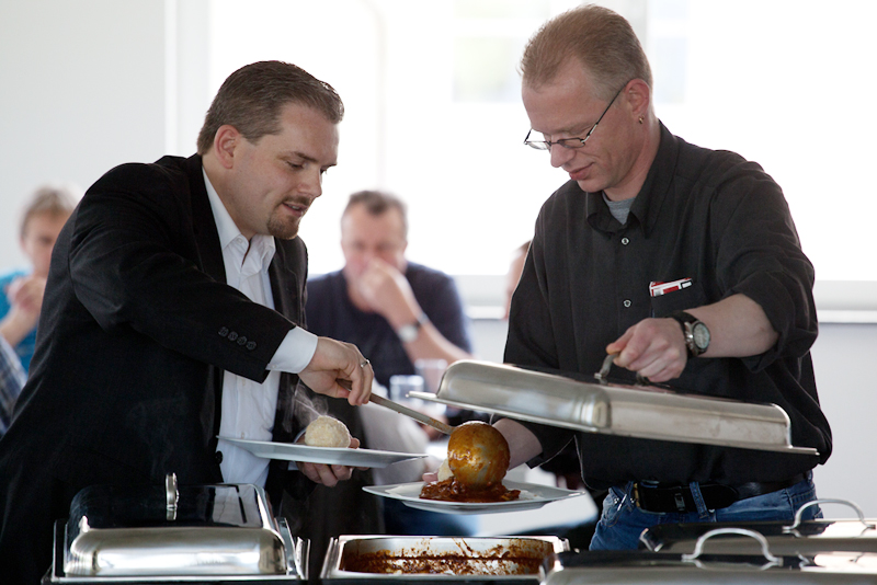 Zwei Männer stehen an einem warmen Buffet, einer hebt den Deckel eines Chafing-Dishes, der andere lässt sich Gulasch mit Kloß auf den Teller geben.