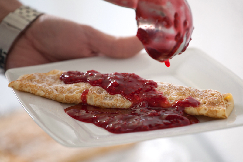 Hand hält einen Teller mit Palatschinken, über den eine rote Fruchtsauce aus einer Kelle gegossen wird.