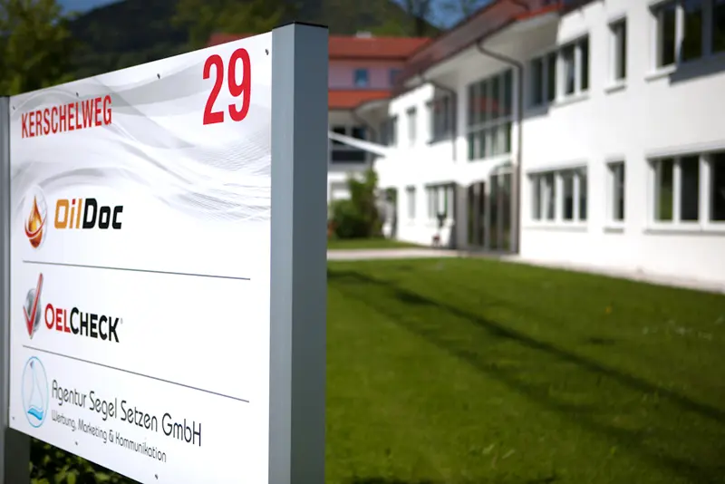 Firmenschild am Kerschelweg 29 mit den Logos von OilDoc, OELCHECK und einer Agentur, im Hintergrund ein weißes Büro- und Schulungsgebäude mit Rasenfläche.