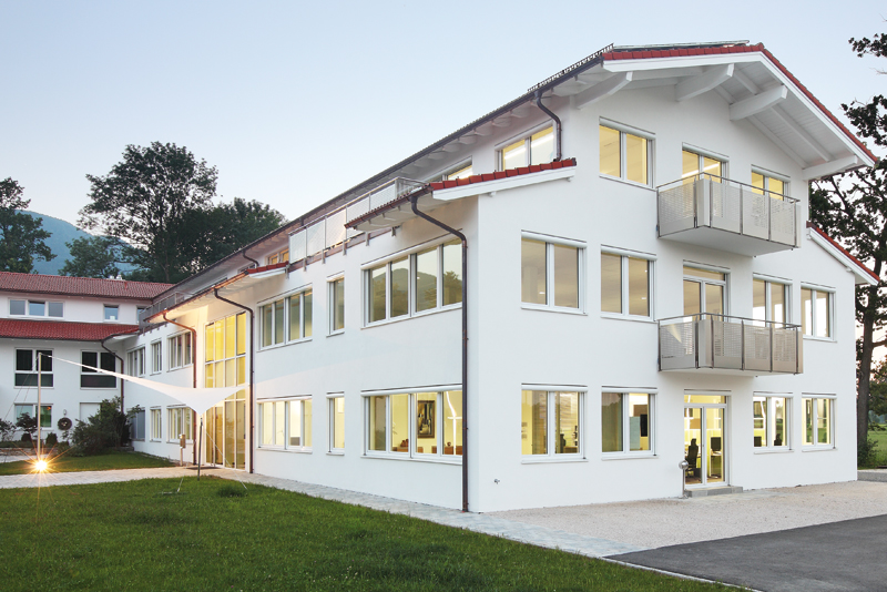 Modernes, weißes Büro- und Schulungsgebäude mit vielen beleuchteten Fenstern und Balkonen in der Dämmerung, umgeben von Rasenfläche und Weg in Brannenburg, Raum Bayern.