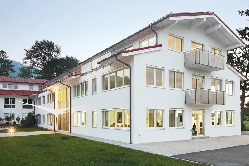 Modernes, weißes Büro- und Schulungsgebäude mit vielen beleuchteten Fenstern und Balkonen in der Dämmerung, umgeben von Rasenfläche und Weg in Brannenburg, Raum Bayern.