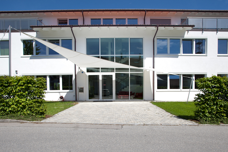 Frontansicht eines modernen weißen Büro- und Schulungsgebäudes mit großflächiger Glasfront und Sonnensegel über dem Haupteingang.