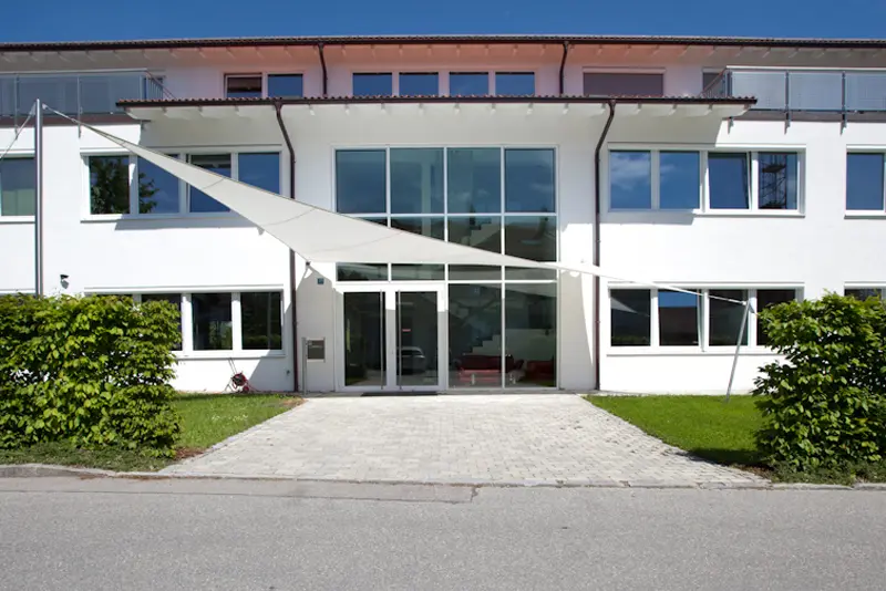 Frontansicht eines modernen weißen Büro- und Schulungsgebäudes mit großflächiger Glasfront und Sonnensegel über dem Haupteingang.