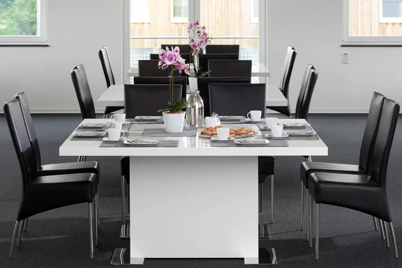 Modern gedeckter Tisch mit schwarzen Stühlen, Tassen, Tellern, Gebäck und einer Kaffeekanne, in der Mitte zwei Orchideen, im Hintergrund weitere Tische im Seminarraum.