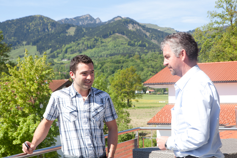 Zwei Seminarteilnehmer unterhalten sich auf einem Balkon vor grünen Hügeln und Bergen, im Vordergrund sind Dächer mit roten Ziegeln zu sehen.