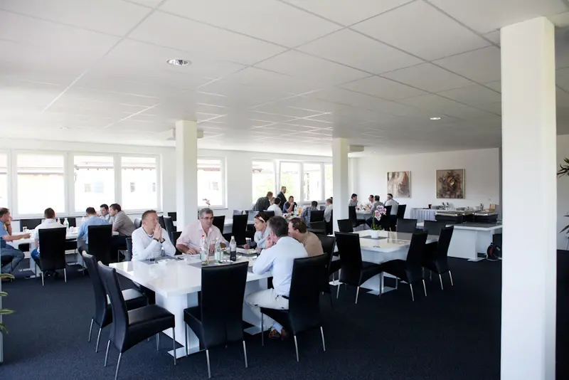 Heller Pausen- und Speiseraum mit mehreren Tischen, an denen Seminarteilnehmer sitzen, essen und sich unterhalten, im Hintergrund ein Buffetbereich.