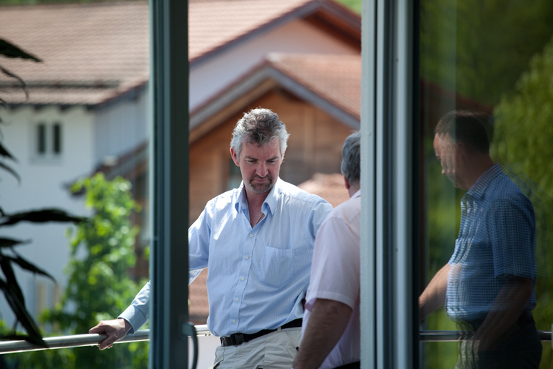 Mehrere Männer im Business-Look unterhalten sich auf einem Balkon vor einem Gebäude mit Satteldach, Spiegelung einer Person in der Glasfront.