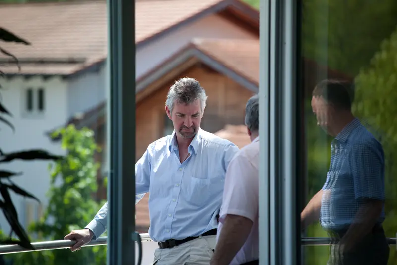 Mehrere Männer im Business-Look unterhalten sich auf einem Balkon vor einem Gebäude mit Satteldach, Spiegelung einer Person in der Glasfront.