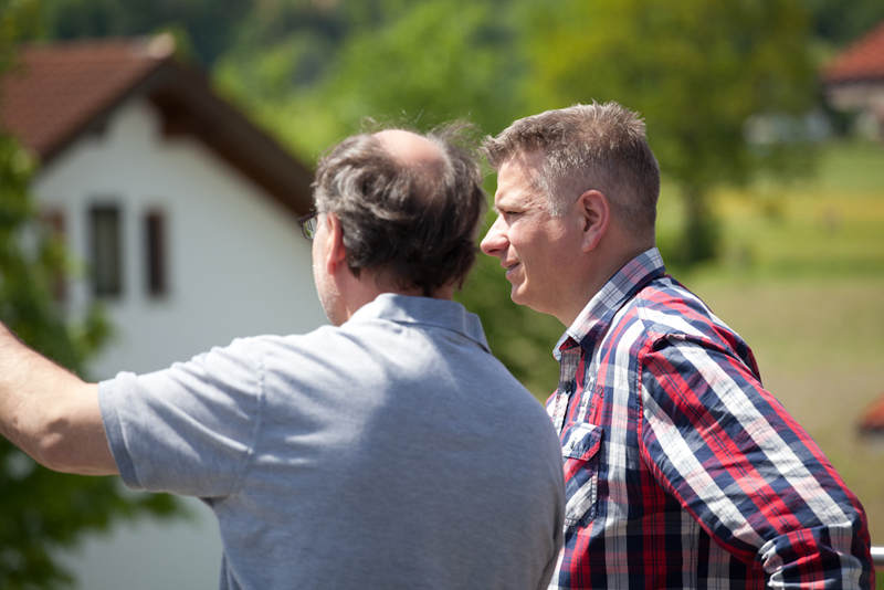 Zwei Männer unterhalten sich im Freien mit Blick auf ein Haus und grüne Umgebung, einer zeigt mit dem Arm in die Landschaft.