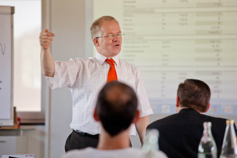 Referent im Seminarraum erklärt vor projizierten Tabellen seine Inhalte, Teilnehmende sitzen ihm zugewandt und hören zu.