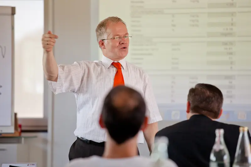 Referent im Seminarraum erklärt vor projizierten Tabellen seine Inhalte, Teilnehmende sitzen ihm zugewandt und hören zu.