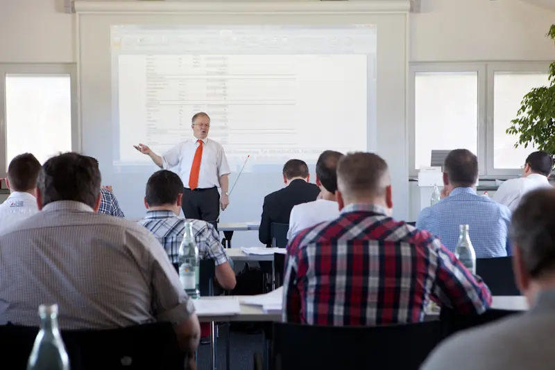 Referent steht vor einer projizierten Präsentation im Seminarraum und erklärt Inhalte, während mehrere Teilnehmende in Reihen zuhören und mitschreiben.