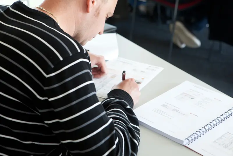 Seminarteilnehmer sitzt am Tisch und füllt konzentriert Arbeitsunterlagen aus, neben ihm liegen Seminar-Skripte und ein aufgeschlagenes Heft.