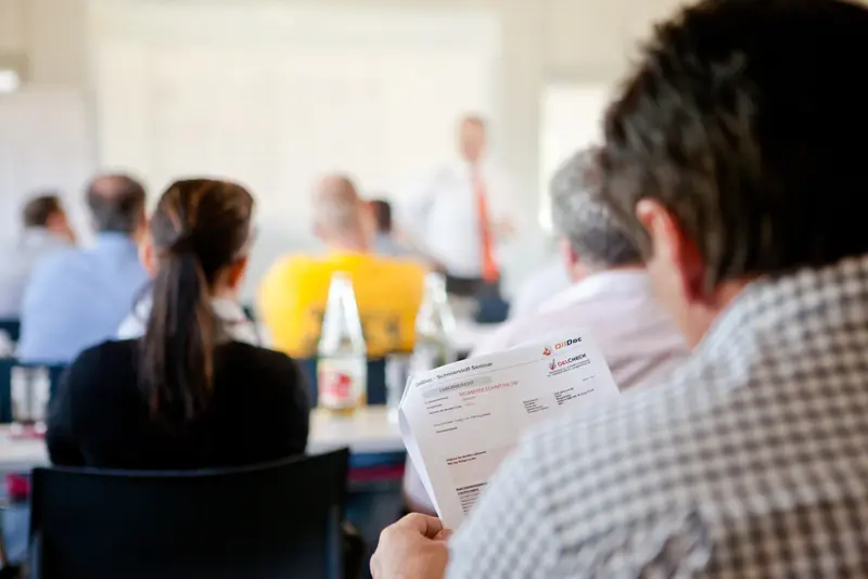 Seminarteilnehmer sitzt im Plenum und liest Schulungsunterlagen, im Hintergrund spricht ein Referent vor der Gruppe.