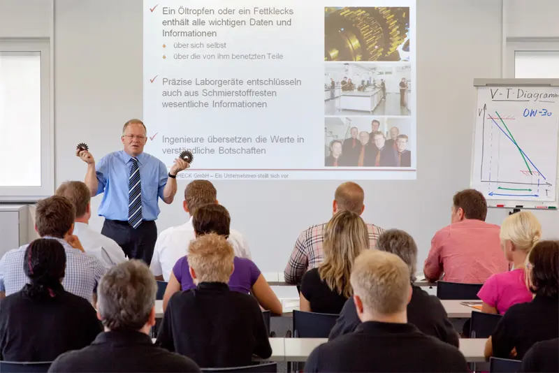 Referent steht vor einer Präsentationsfolie zu Öltropfen, Laboranalyse und Ingenieurleistungen und erklärt einer Gruppe von Teilnehmenden die Inhalte.
