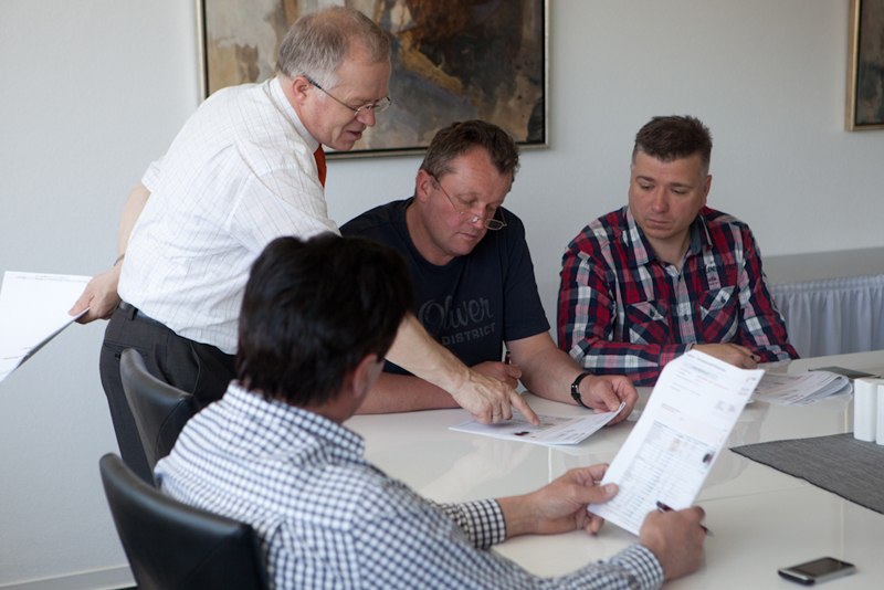 Mehrere Männer sitzen an einem Tisch und besprechen gemeinsam Unterlagen, ein Moderator zeigt auf eine Stelle im Dokument und erklärt etwas.