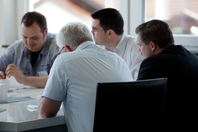 Vier Männer sitzen in einer Kleingruppe an einem Tisch und arbeiten konzentriert an Unterlagen in einem hellen Raum mit großen Fenstern.