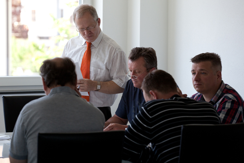 Dozent steht an einem Tisch und unterstützt eine Kleingruppe von Teilnehmern, die gemeinsam an Seminarunterlagen arbeiten.