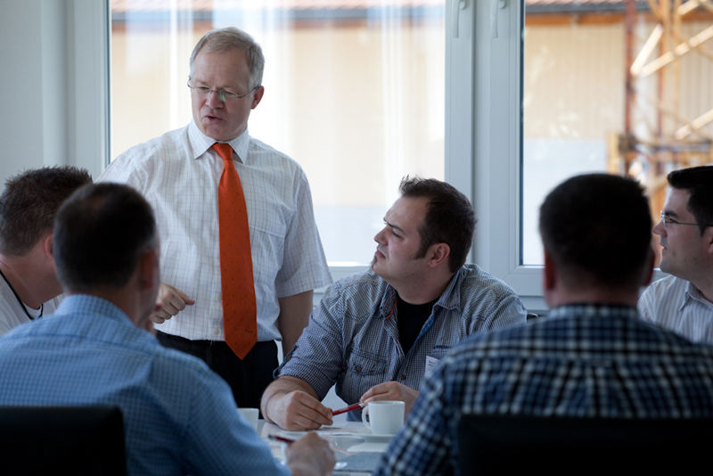 Dozent im weißen Hemd mit orangefarbener Krawatte spricht mit einer Kleingruppe von Seminarteilnehmern, die an einem Tisch im hellen Raum sitzen.