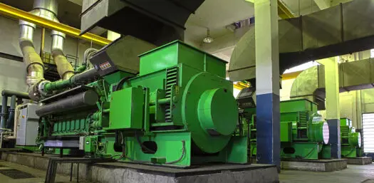 Mehrere große grüne stationäre Gasmotoren in einem gut beleuchteten Maschinenraum mit Rohrleitungen und Abluftsystemen