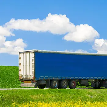 Blauer Sattelzug fährt bei sonnigem Wetter über eine Landstraße vor grünen Feldern