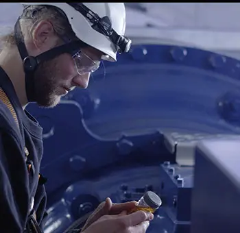 Servicetechniker mit Helm und Schutzbrille prüft eine Ölprobe vor dem großen Getriebe einer Windkraftanlage im Maschinenhaus