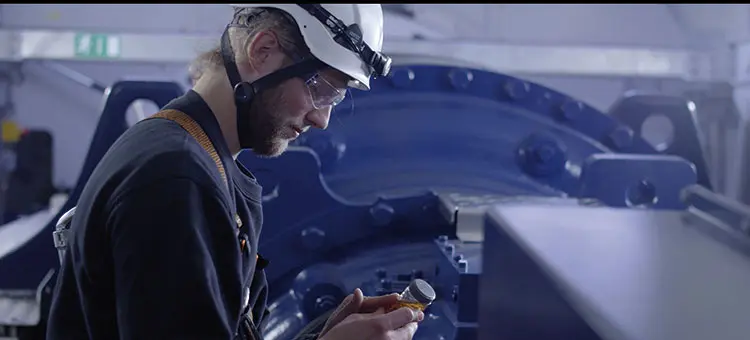 Servicetechniker mit Helm und Schutzbrille prüft eine Ölprobe vor dem großen Getriebe einer Windkraftanlage im Maschinenhaus